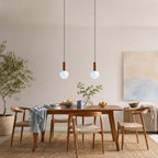 Two Clérachic pendants with pale blue globes and wood stems hang over a timber dining table, with a soft abstract painting behind.