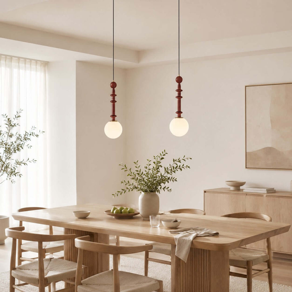 A pair of white-globe Clérachic pendants hangs over a light wood dining table with woven-seat chairs and a plant.