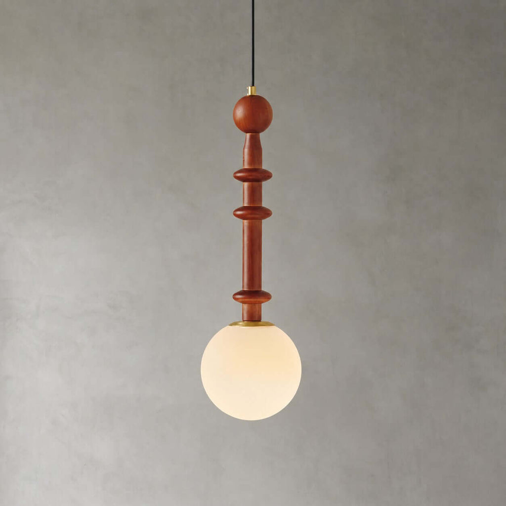This Clérachic pendant has a slim turned wooden stem and a frosted white globe, lit warmly against a grey wall.