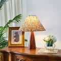 Floral-shaded Clérachic table lamp on a carved wooden sideboard, beside a framed bear print, a green plant and a gold basket of white flowers.