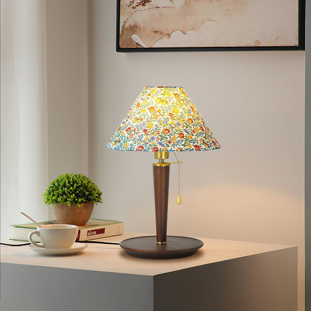 Clérachic-listed table lamp with a floral conical shade on a slim dark column and round tray base, a pull cord hanging, beside a cup and a small plant.