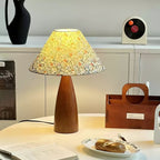 Lit Clérachic table lamp on a white table, its floral shade glowing yellow, next to a wooden organiser box and a bread roll on a plate.