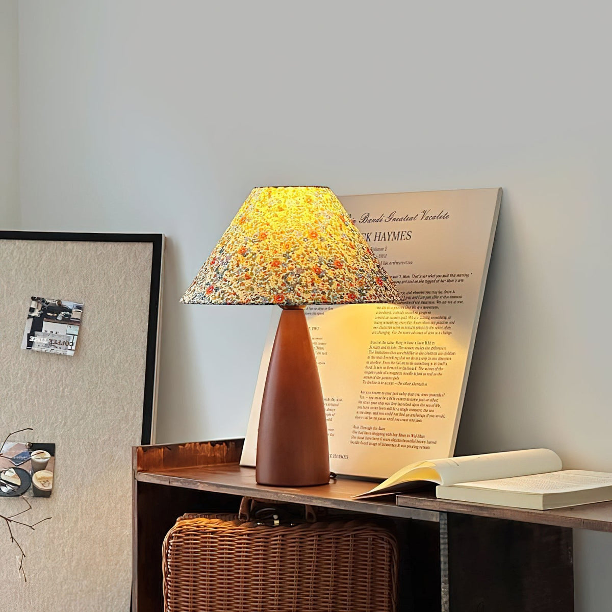 Glowing Clérachic table lamp on a low shelf in front of a printed poster, with a woven basket below and an open book beside it.