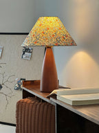 Clérachic table lamp lit warm on a dark wooden shelf, the floral shade glowing, with a corkboard behind and a basket underneath.