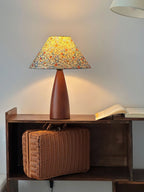 Upright view of this Clérachic table lamp lit above a wicker case on a dark shelf, with a warm pool of light on the grey wall.