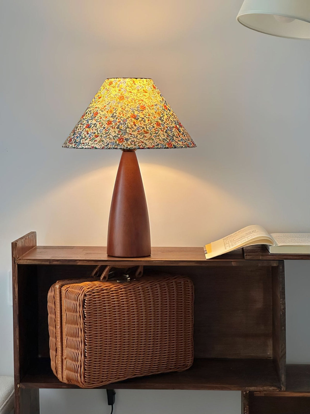 Upright view of this Clérachic table lamp lit above a wicker case on a dark shelf, with a warm pool of light on the grey wall.