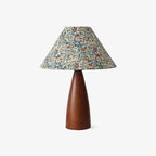 Front view of the Clérachic table lamp, with a blue, red and cream floral shade over a smooth brown tapered wooden base, on white.