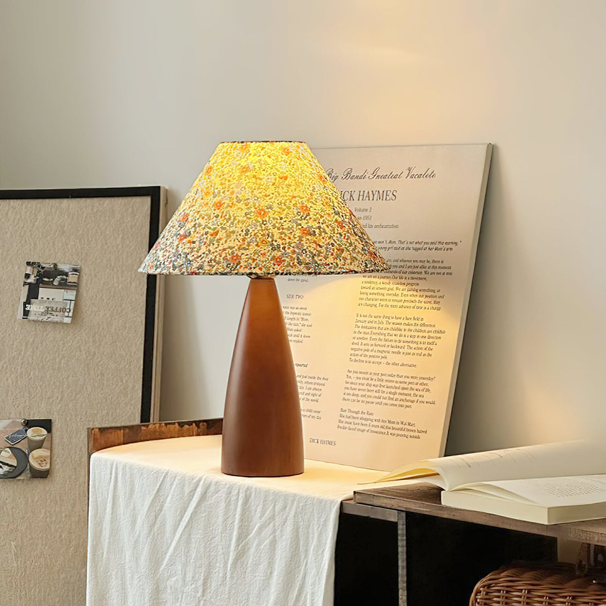 Clérachic table lamp lit on a draped white cloth, the floral shade glowing yellow, in front of a printed poster and an open book.