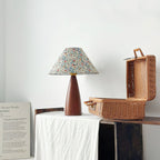 Small Clérachic table lamp on a white cloth next to an open wicker picnic basket, with a printed poster leaning against the wall.