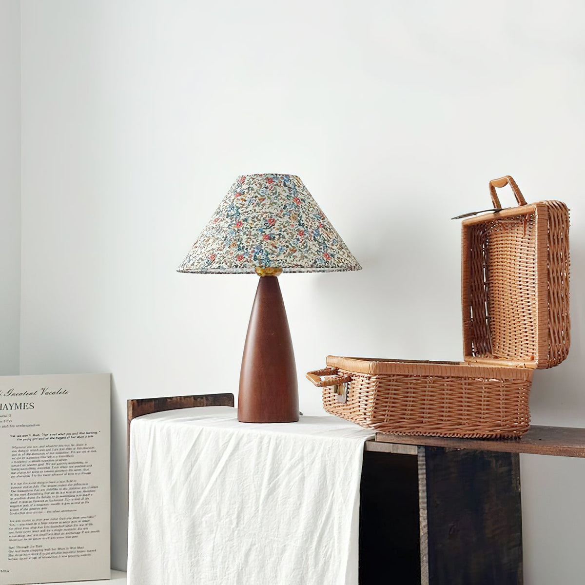 Small Clérachic table lamp on a white cloth next to an open wicker picnic basket, with a printed poster leaning against the wall.