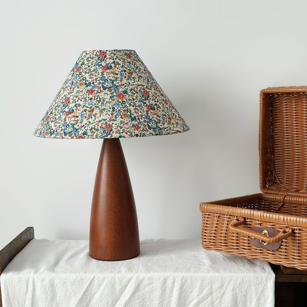 Clérachic table lamp with a floral shade and wooden base on a white cloth, beside an open wicker basket, against a pale wall.