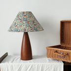Clérachic table lamp with a floral shade and wooden base on a white cloth, beside an open wicker basket, against a pale wall.