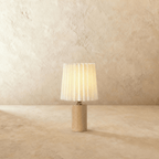 Clérachic table lamp with a pleated cream shade glowing warm above a slim light oak cylinder base on a plaster-toned backdrop.