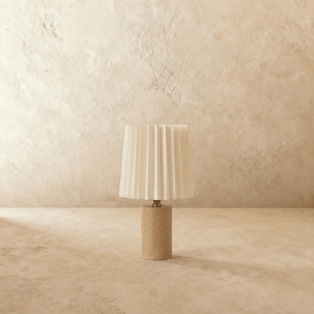 Clérachic table lamp with an unlit pleated cream shade above a light natural wood cylinder base, brass-toned neck, plaster backdrop.