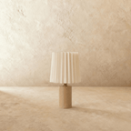 Clérachic table lamp with an unlit pleated cream shade above a light natural wood cylinder base, brass-toned neck, plaster backdrop.