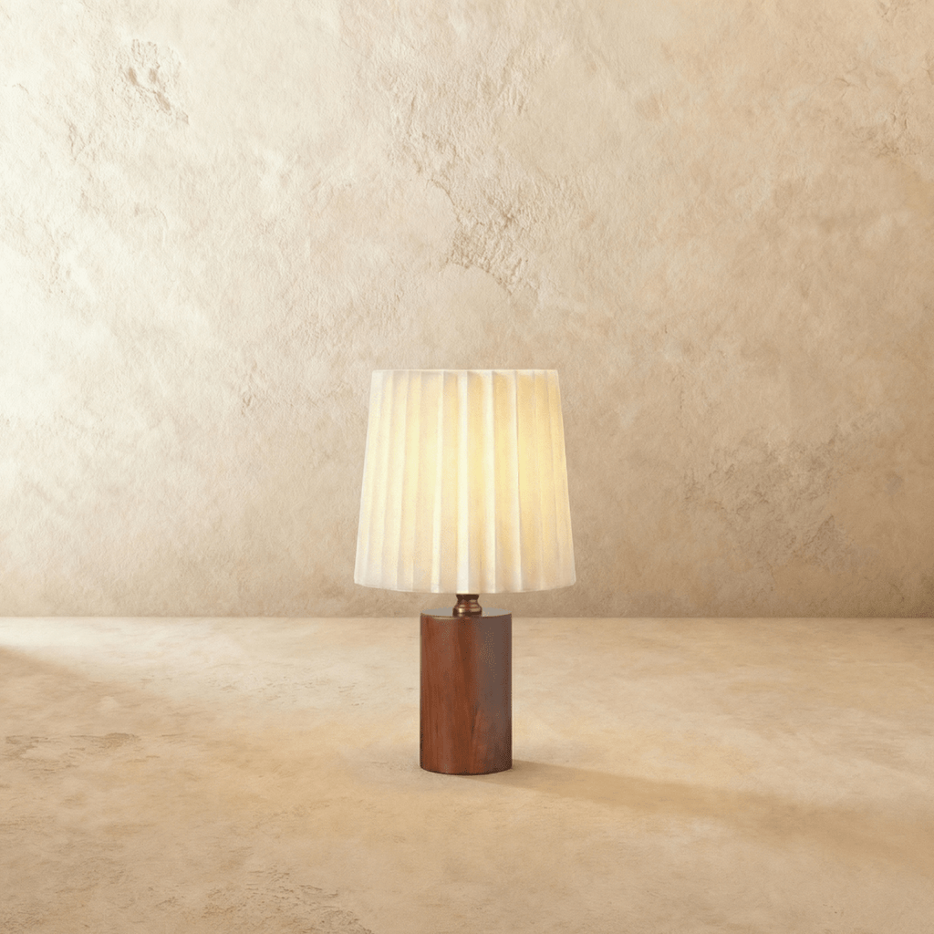 Clérachic table lamp with a glowing pleated cream shade above a dark walnut cylinder base on a plaster-toned backdrop.