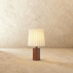 Clérachic table lamp with a glowing pleated cream shade above a dark walnut cylinder base on a plaster-toned backdrop.