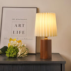 Clérachic table lamp with a glowing pleated shade and walnut cylinder base on a dark sideboard beside a framed print and flowers.