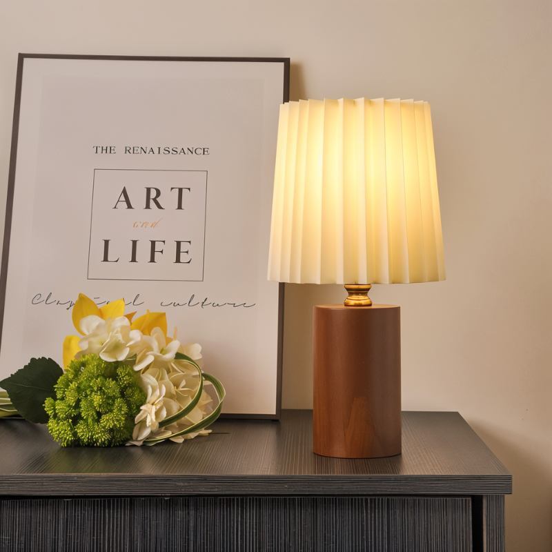 Clérachic table lamp with a glowing pleated shade and walnut cylinder base on a dark sideboard beside a framed print and flowers.