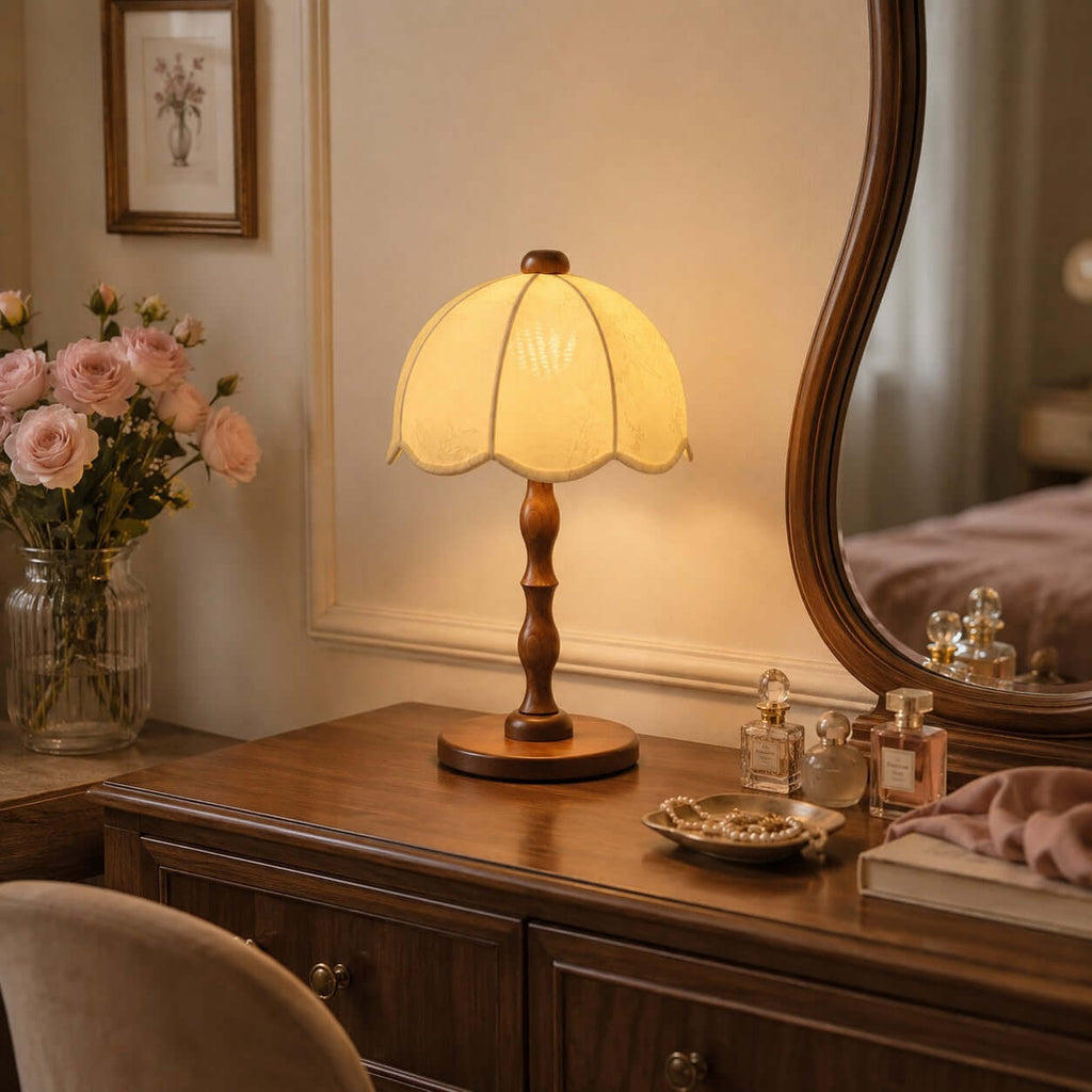 A Clérachic table lamp lit on a wooden dresser, beside a curved mirror, a vase of pink roses and small perfume bottles.
