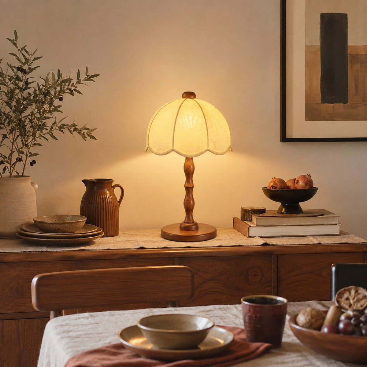 A Clérachic table lamp lit on a sideboard with ceramic jugs, a fruit bowl and a stack of books, beside a dining table.