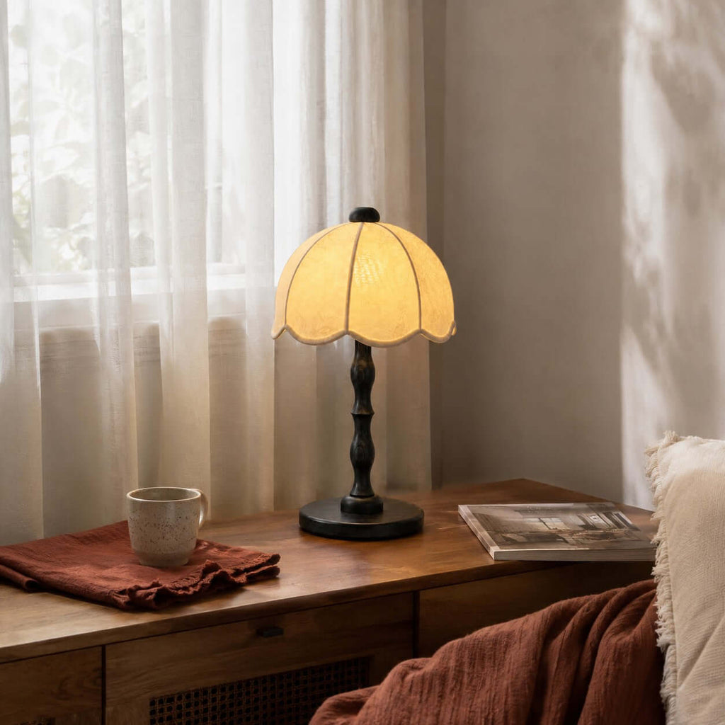 A Clérachic table lamp with a black base and scalloped shade, on a low wooden cabinet by sheer curtains and a rust throw.
