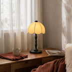 A Clérachic table lamp with a black base and scalloped shade, on a low wooden cabinet by sheer curtains and a rust throw.