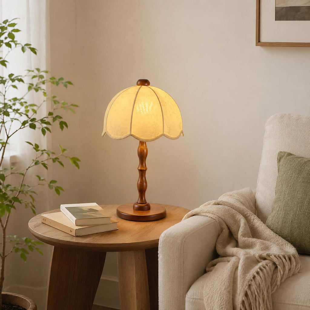 Clérachic table lamp with a walnut stem on a round wooden side table beside a cream armchair, a green cushion and a throw.