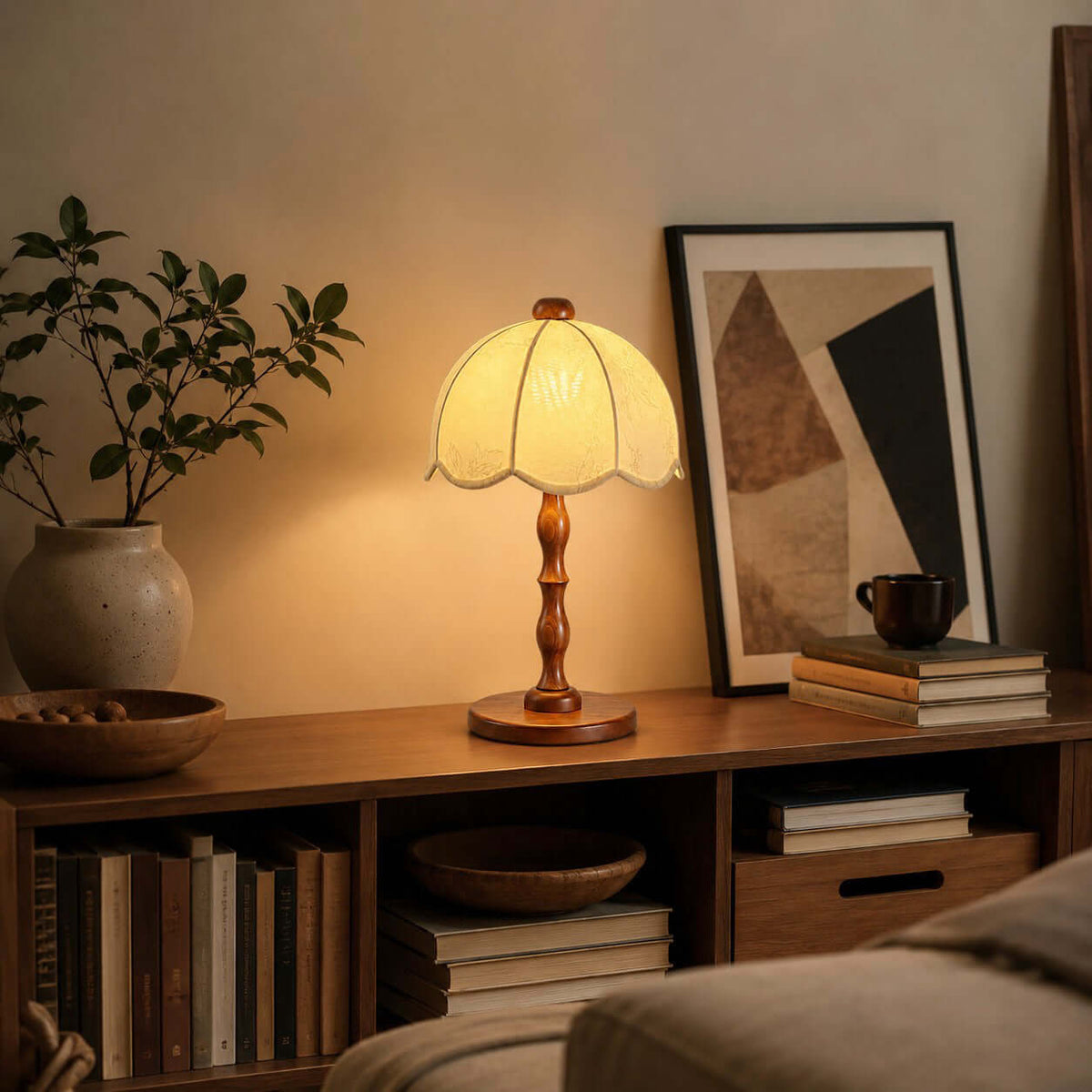 Scalloped-shade Clérachic table lamp lit on a low bookshelf, with a framed print, a vase and a ceramic bowl.