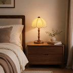 This Clérachic table lamp lit on a walnut bedside table next to a bed with cream bedding and a small plant in a vase.