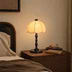 Black-based Clérachic table lamp with a scalloped shade on a bedside table, with a cup and a book on a small tray.