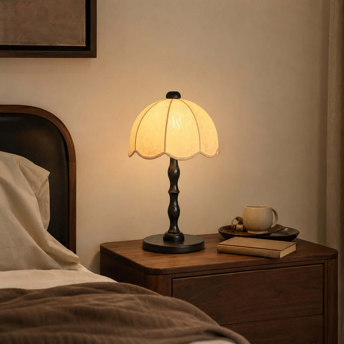 Black-based Clérachic table lamp with a scalloped shade on a bedside table, with a cup and a book on a small tray.