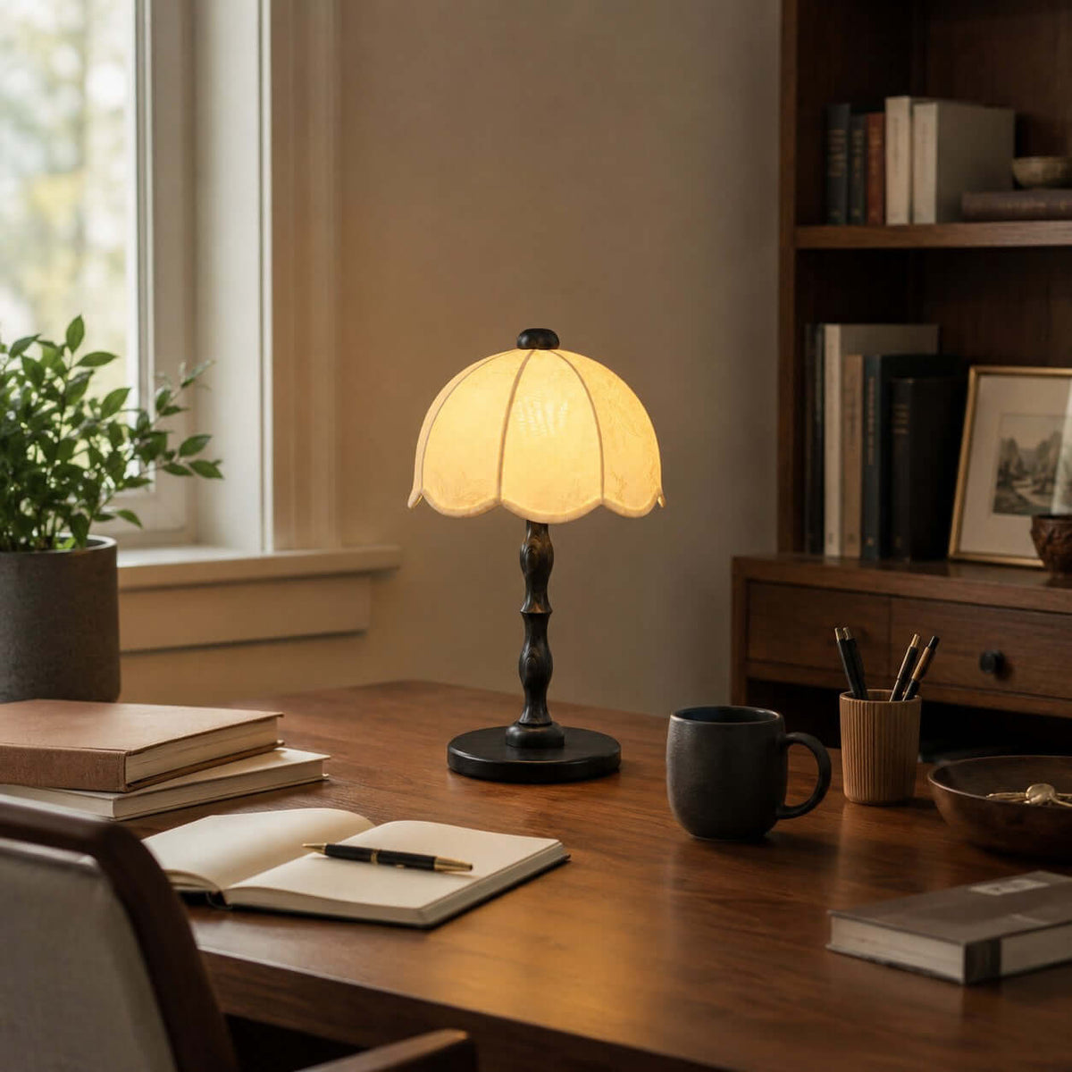 A Clérachic table lamp with a black base on a wooden desk by a window, with an open notebook, a mug and a pen pot.