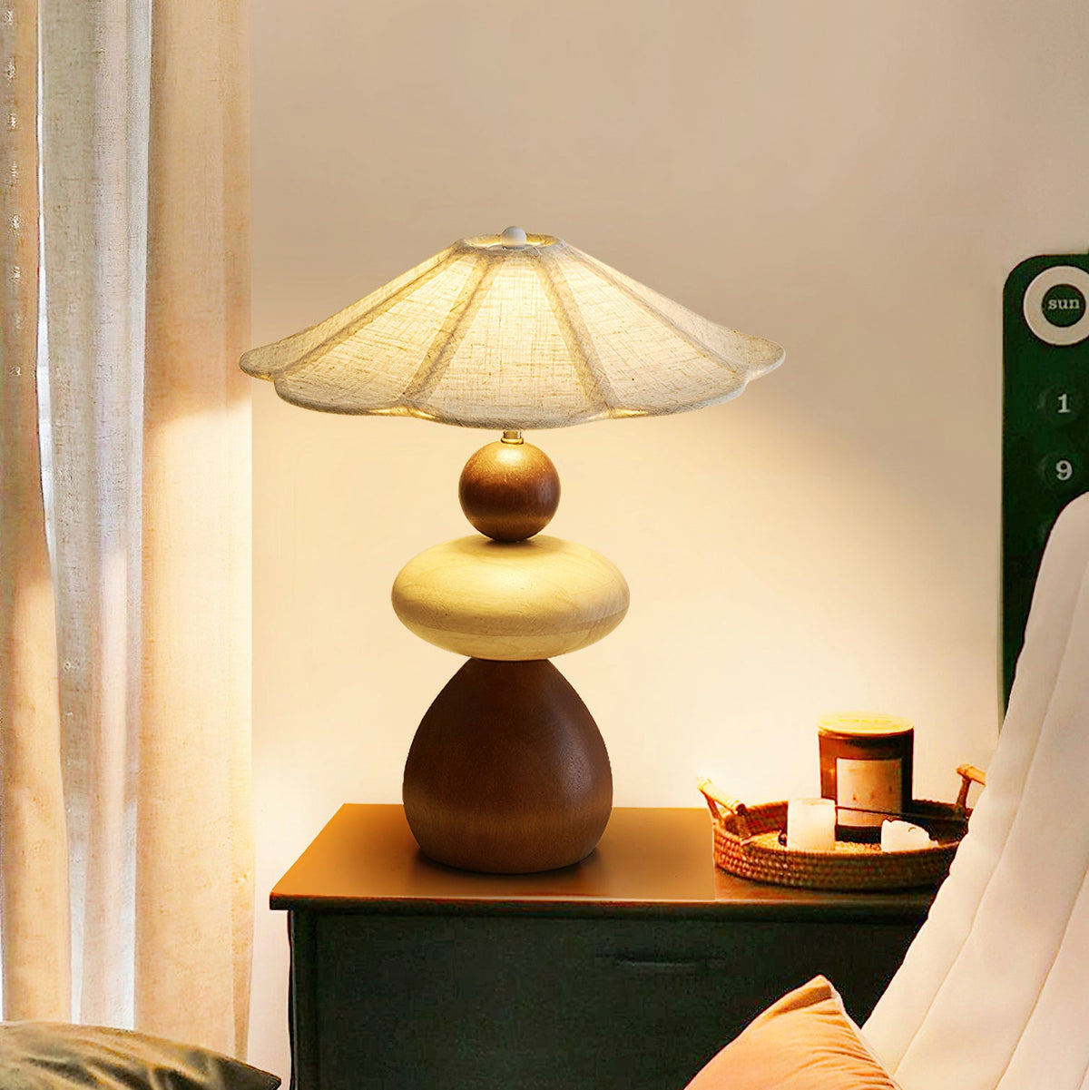 Lit Clérachic table lamp on a dark green bedside chest, with a wicker tray, a candle and a pale curtain behind it.