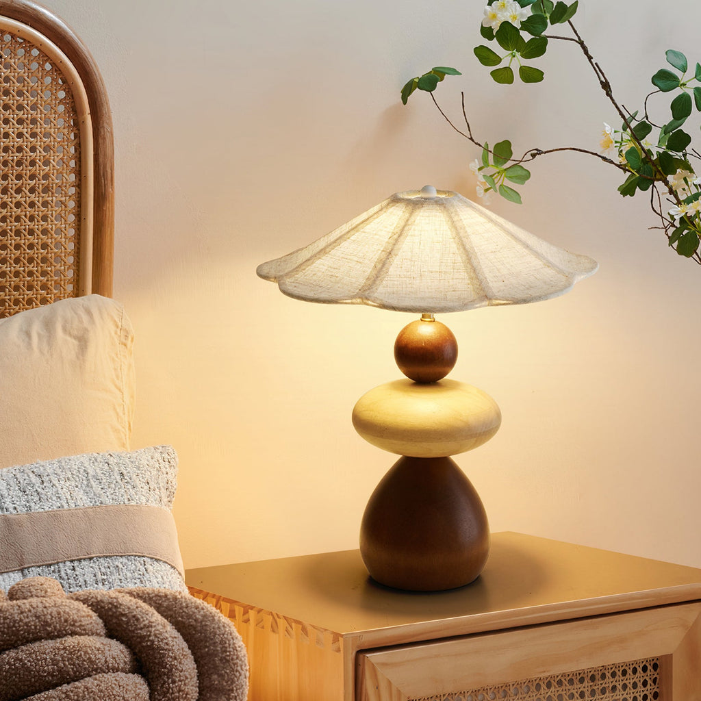 A Clérachic table lamp lit beside a cane-fronted bedside table, with a knitted throw and a flowering branch in the background.