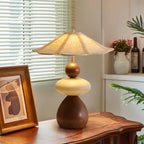 A Clérachic table lamp on a wooden console, with a framed print in the foreground and a vase of pink flowers by the window.