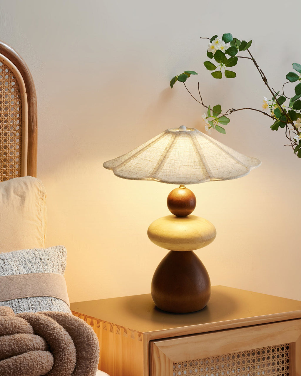 A Clérachic table lamp on a cane-fronted bedside table, with a pale cushion, a chunky knit throw and a leafy branch.