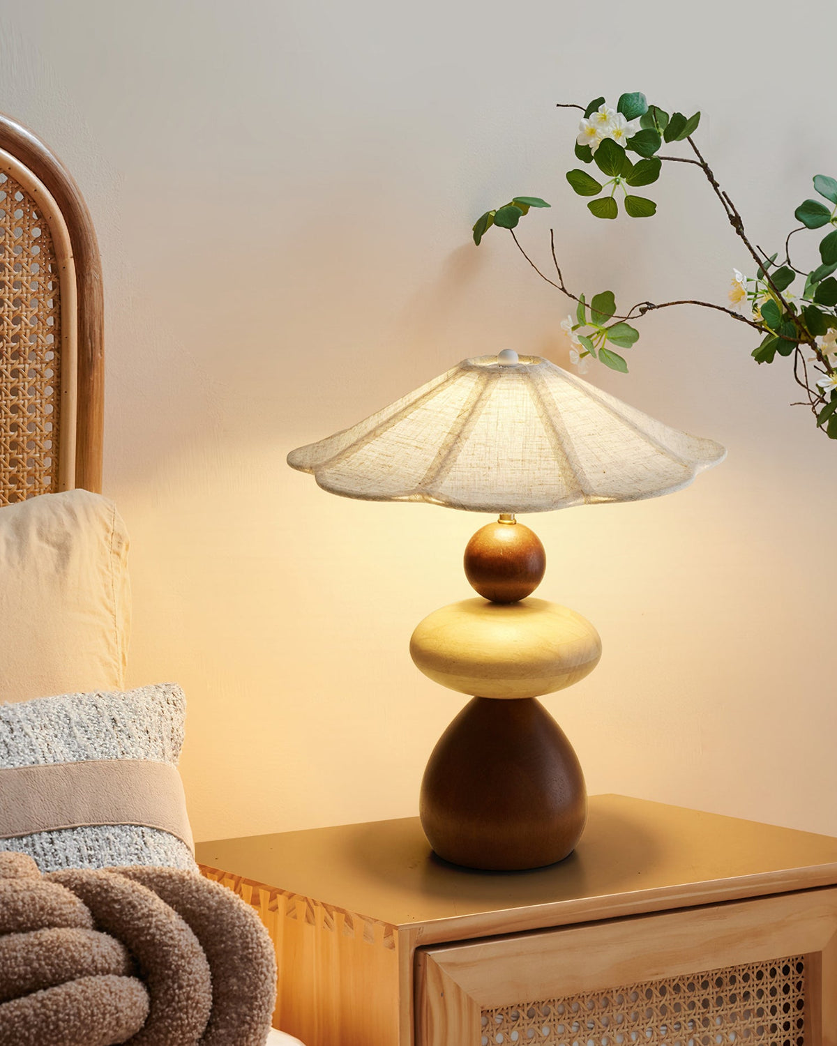 A Clérachic table lamp on a cane-fronted bedside table, with a pale cushion, a chunky knit throw and a leafy branch.