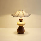 A Clérachic table lamp glowing warmly on a pale tabletop against a soft peach backdrop, its flared linen shade above the wood body.