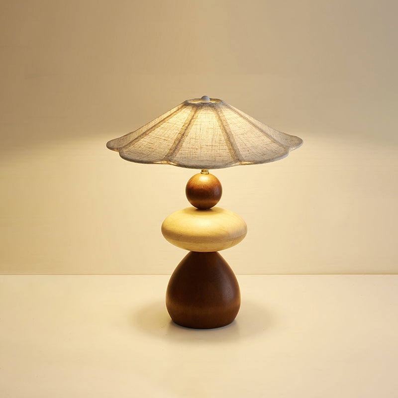 A Clérachic table lamp glowing warmly on a pale tabletop against a soft peach backdrop, its flared linen shade above the wood body.