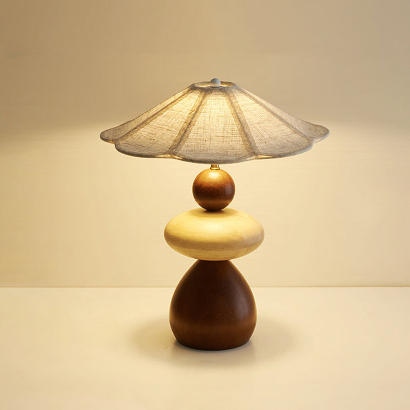 Front view of this Clérachic table lamp lit on a cream surface, with its flared linen shade and stacked walnut and pale wood body.