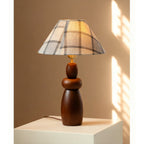 Sun falls across a white plinth holding a Clérachic table lamp with a plaid fabric shade and a stacked, turned wood base.