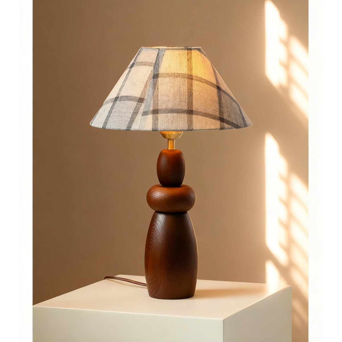 Sun falls across a white plinth holding a Clérachic table lamp with a plaid fabric shade and a stacked, turned wood base.