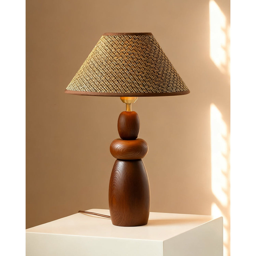 A Clérachic table lamp with a straw weave shade and a stacked wood base, standing on a white plinth against a warm tan wall.