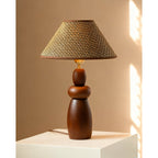 A Clérachic table lamp with a straw weave shade and a stacked wood base, standing on a white plinth against a warm tan wall.