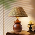 A Clérachic straw weave shade lamp on a rounded wood base glows on a sideboard, beside a candle holder and a striped blind.
