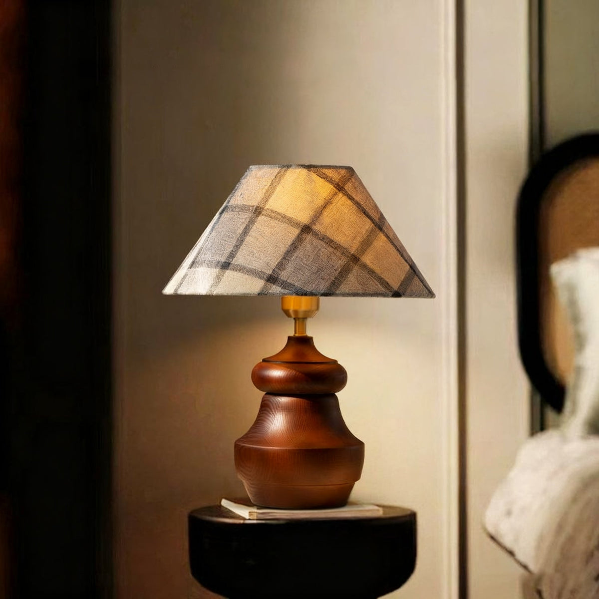 A Clérachic plaid-shade table lamp with a wood base stands on a small black table, with a book below and a rattan chair behind.