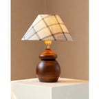 In a corner of a tan wall, a Clérachic table lamp with a plaid shade and a rounded wood base stands on a white plinth.