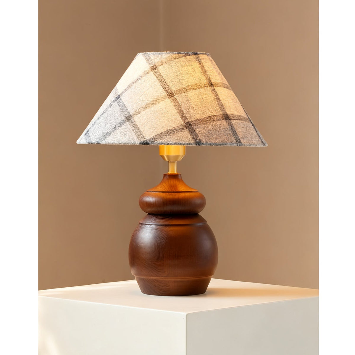 In a corner of a tan wall, a Clérachic table lamp with a plaid shade and a rounded wood base stands on a white plinth.