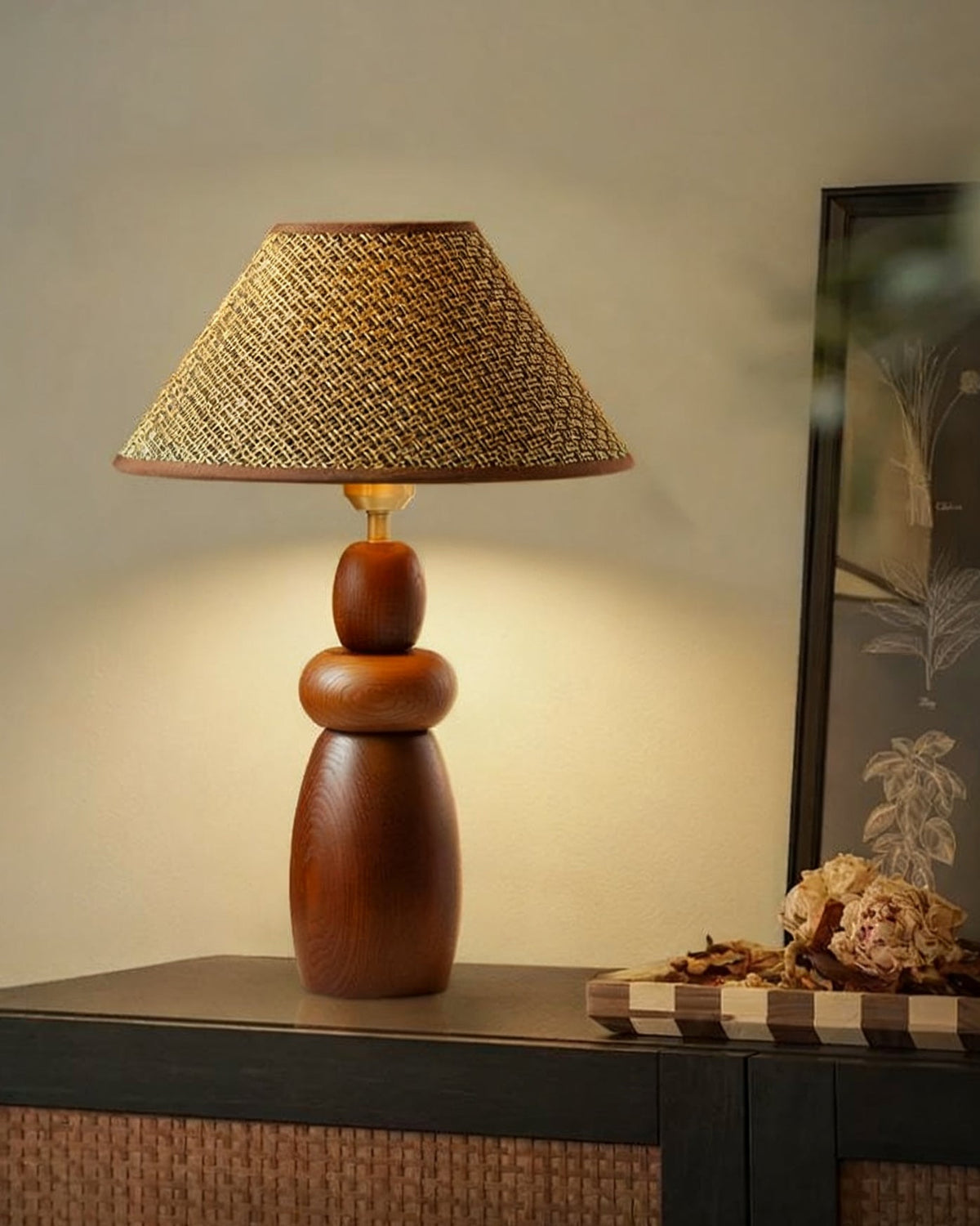 A Clérachic straw weave shade lamp on a tall stacked wood base stands on a dark rattan cabinet, beside a framed print.